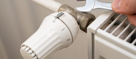 Detail termostatické hlavice a kovového spoje radiátoru, nad kterým člověk drží otevřený klíč