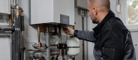 Servisní technik kontroluje nástěnný plynový kotel v technické místnosti. Pod kotlem je vidět potrubí, ventily a manometr